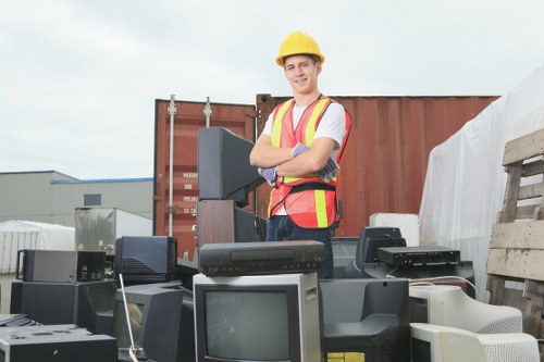 A man wearing a yellow safety helmet and a high-visibility vest stands outdoors at a waste removal site surrounded by discarded electronic equipment, including CRT monitors, desktop computers, keyboards, and other obsolete technology. The electronic waste is arranged in a cluttered manner on a paved surface, with additional materials and pallets visible in the background. He is smiling and has his arms crossed, indicating confidence in his work. The setting appears to be an industrial or waste collection area, with a shipping container and warehouse structures in the distance. Natural daylight illuminates the scene, highlighting the contrast between the old, bulky technology and the safety gear. This image relates to rubbish removal services provided by Office Clearance Bermondsey, supporting efficient clearance of electronic waste in an urban environment like Bermondsey, London, or nearby areas.