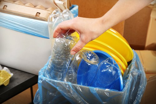 A close-up view of a person's hand holding a clear plastic bottle filled with water, positioned above a black surface that supports a large open-top container lined with blue plastic. Inside the container are multiple plastic buckets in yellow and blue colours, stacked and leaning against each other. The scene appears to be indoors, likely in an office or storage area, with neutral brown and beige tones visible in the background. The arrangement suggests an action related to waste or rubbish collection, consistent with rubbish removal services offered by Office Clearance Bermondsey, as seen in the website context. The focus is on the plastic waste items and their handling, highlighting the materials' textures and colours, with a clean and organized environment for rubbish disposal or recycling activities in a Bermondsey setting.