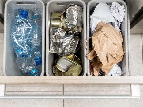 The image displays three grey plastic recycling bins placed side by side on a tiled floor, likely outdoors or in a commercial area. The left bin contains several clear plastic bottles, some with blue caps, crumpled and empty. The middle bin is filled with used, unlabelled metal cans, including some crushed and overlapping, with smooth, reflective surfaces and edges typical of food or beverage containers. The right bin holds a mix of crumpled paper and brown kraft paper, with some white tissue or paper towel waste on top, appearing loosely stacked and slightly messy. The setting appears clean and organized, possibly part of a waste management or rubbish collection setup, relevant to office clearance or general waste disposal services. This depiction is fitting for a professional rubbish removal provider like Office Clearance Bermondsey, serving areas including Bermondsey and nearby postcode regions, highlighting typical waste materials handled in their service categories.