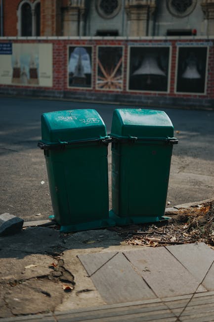 The image depicts a row of three large waste dumpsters positioned along a city sidewalk in front of a building with a purple brick wall. The two black dumpsters are fitted with wheels for mobility and