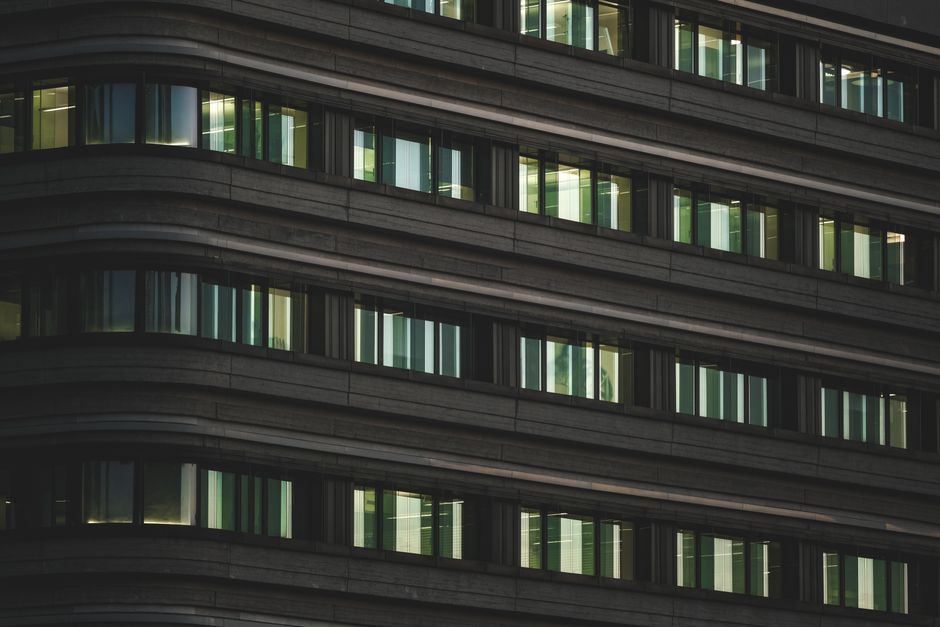 A modern multi-storey office building with a facade composed predominantly of large, reflective glass windows set within dark framing. The building's exterior features horizontal bands of black metal 