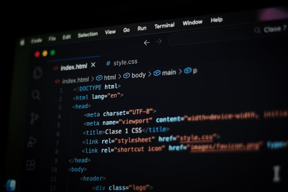 A close-up view of an open silver laptop with a black keyboard displaying lines of colorful computer code on a dark screen, positioned on a light wooden desk. To the left of the laptop, there is a whi